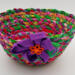This vibrant multi-colored fabric rope bowl is ready for multiple uses. This view shows the purple flower with orange button. This vibrant multi-colored fabric rope bowl is ready for multiple uses. This view shows the purple flower with orange button.